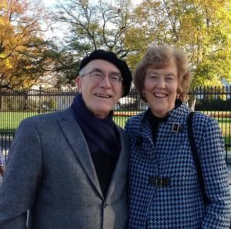man and woman pictured together outside with background of trees with leaves changing colors. Man is wearing a black hat, black shirt and gray blazer. Women is wearing gray blazer.